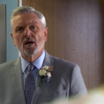 Thomas 'Rocky' Cotton in a gray classic suit with flowers on it standing near the door and looking forward