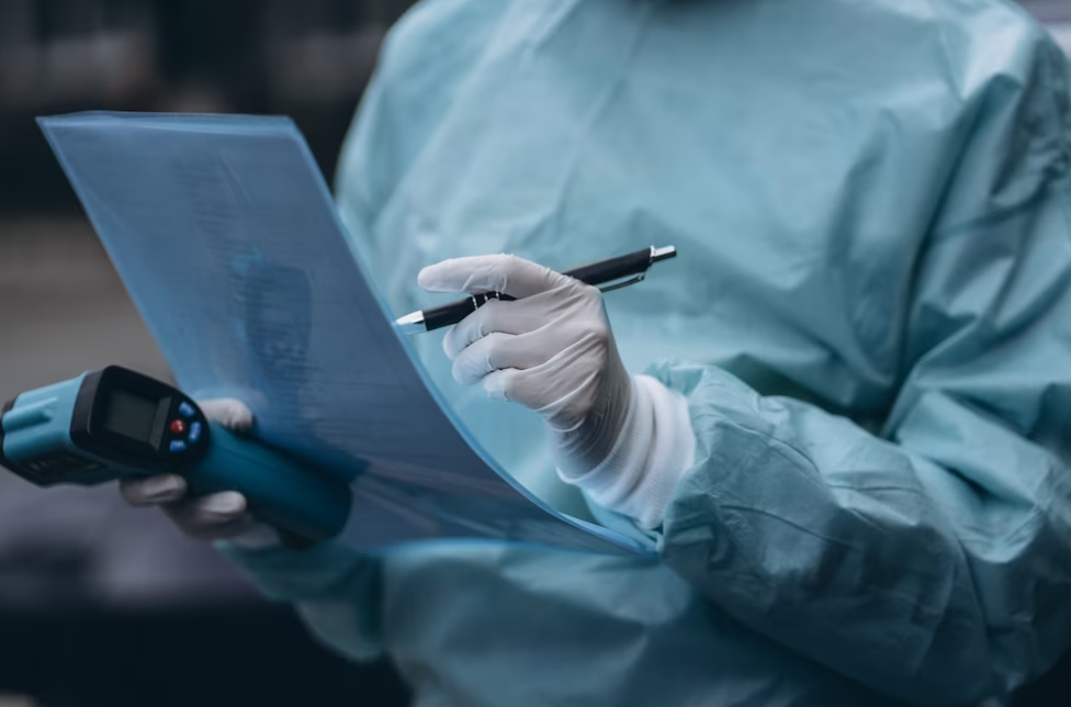 person in blue medical clothing and white gloves holding papers and pen in one hand, and thermometer in another